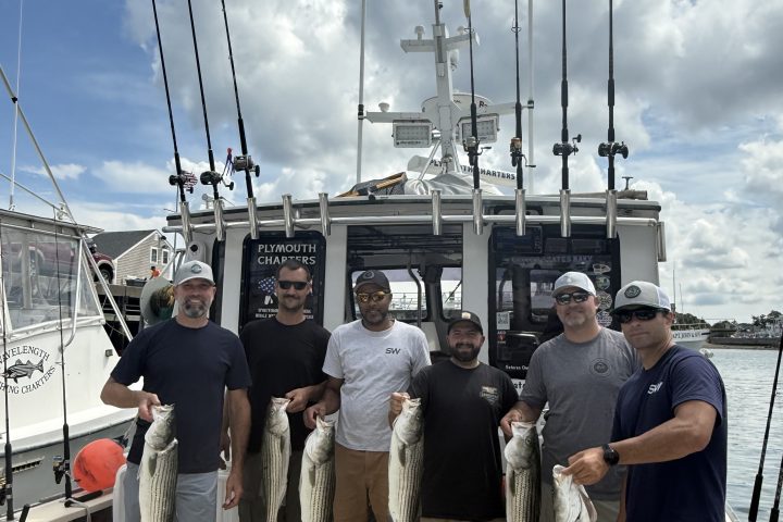 Six men on a boat holding fish, with fishing rods and an American flag in the background.
