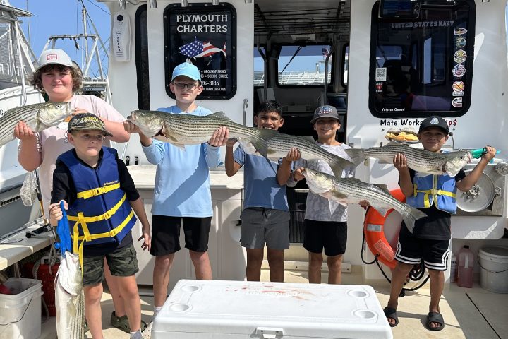 Five kids holding fish in front of a boat with fishing gear and 'Plymouth Charters' sign in the background.
