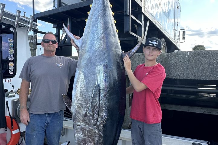Two people standing next to a large tuna hanging by its tail on a fishing boat.