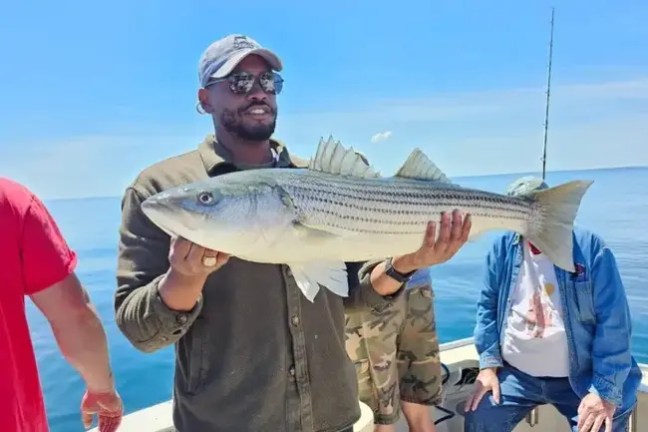 Man holding a large fish on a boat with a fishing rod and sea in the background.
