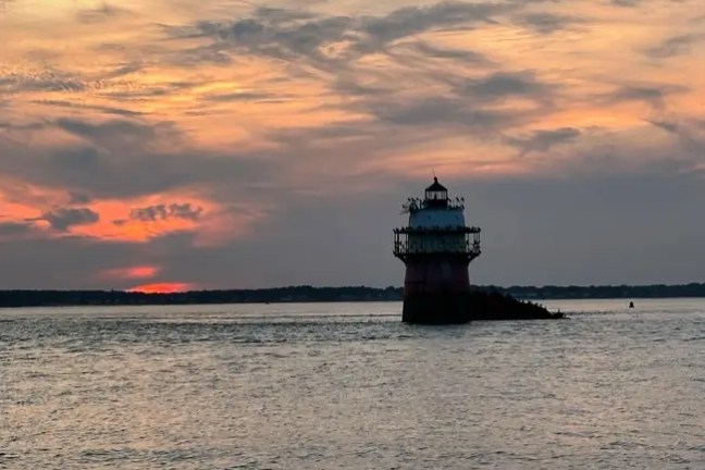 Lighthouse at sunset on calm water with a colorful sky.