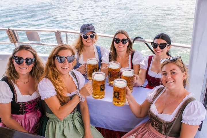 Six women in traditional dresses holding beer mugs on a boat.