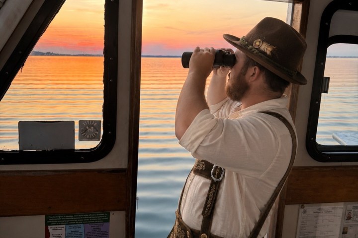 Man in traditional German attire using binoculars on a boat at sunset.